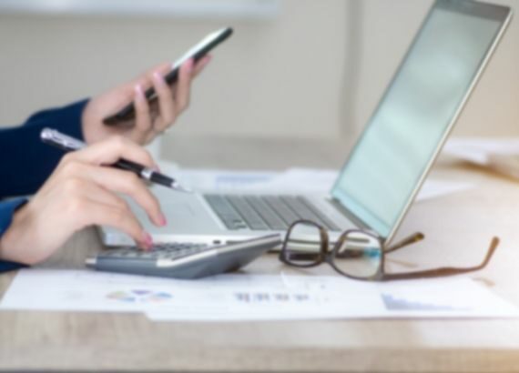 Hände einer Person am Laptop mit Handy und Stift in der Hand